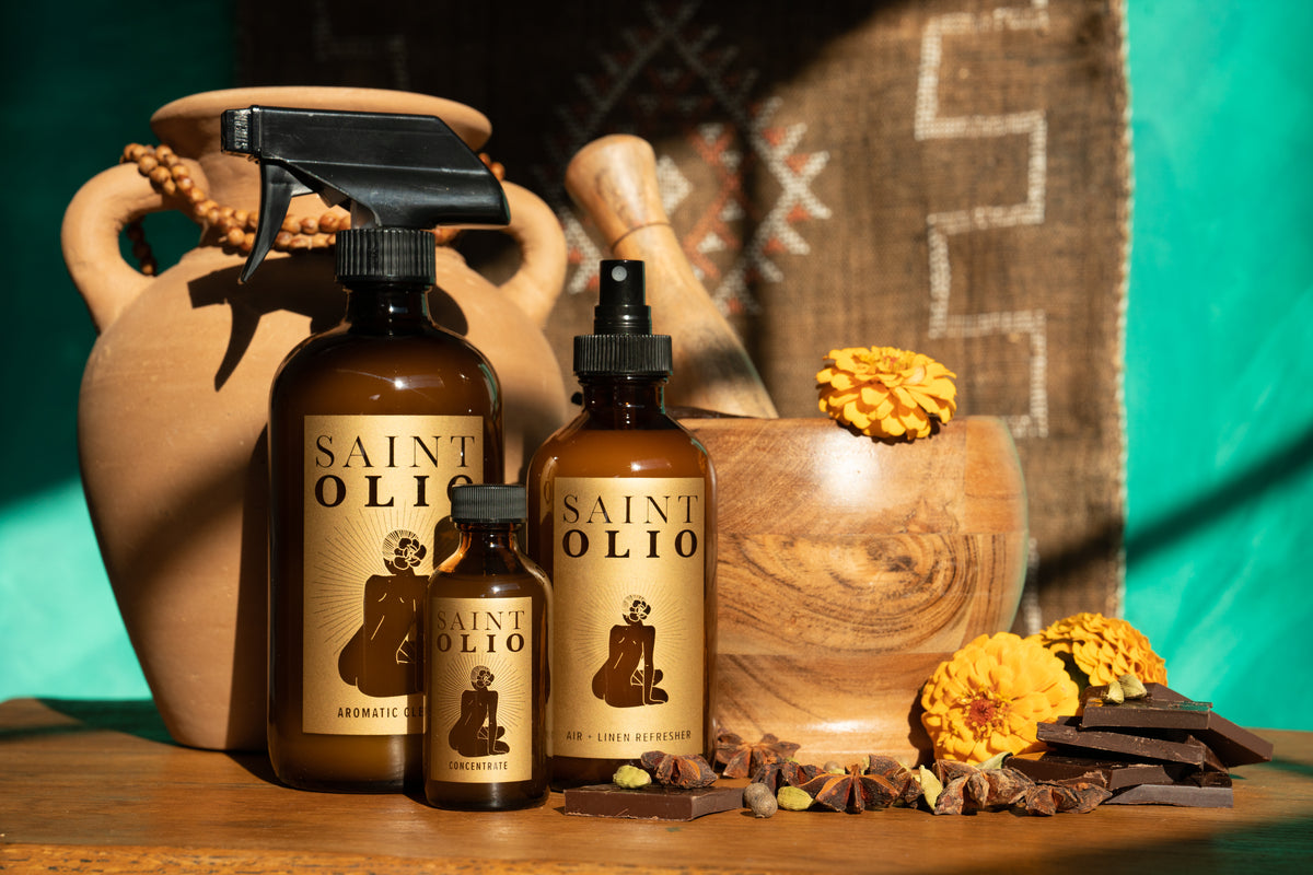 Three Saint Olio essential oil aromatic cleaner bottles in amber glass with black sprayers, styled with a terracotta vase, wooden mortar and pestle, yellow marigolds, and chocolate pieces on a wooden surface.